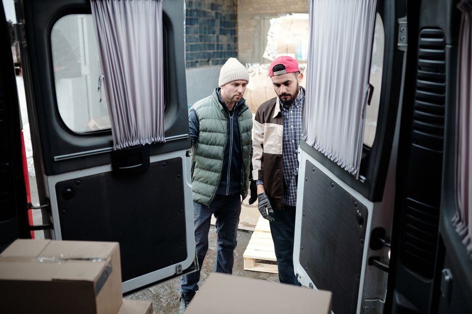 A man wearing a black padded vest, blue long-sleeve shirt, black trousers, and white footwear stands next to an open van door during a home relocation process, with his right hand on his hip and looki